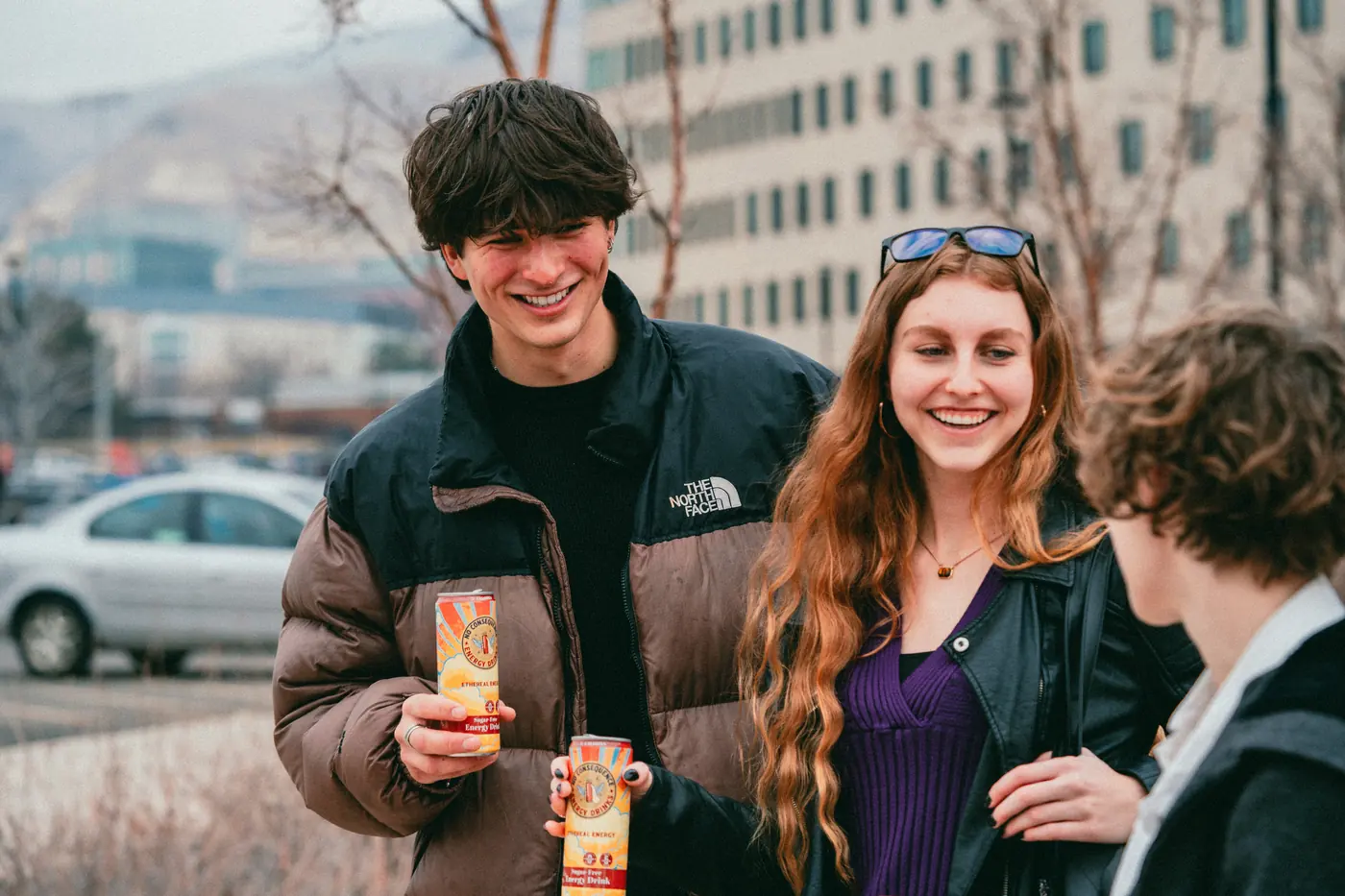 Two friends laughing outdoors in winter, each holding a can of No Consequence energy drink